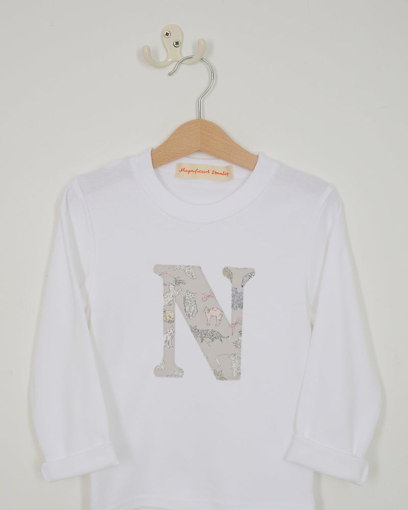 A white long-sleeve t-shirt for kids, hanging on a wooden hanger. The shirt features a large decorative letter 'N' on the front filled with a Liberty Koh & Eito cats print in muted gray, beige, and pink tones.