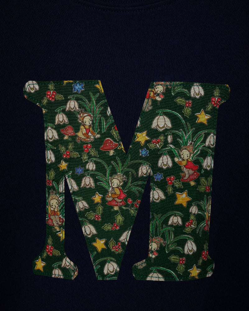 A close-up of a dark navy blue cotton kids t-shirt featuring a large letter 'M' printed on the front, filled with a detailed Liberty woodland fabric of green leaves, white snowdrop flowers, red mushrooms, elves and yellow stars.