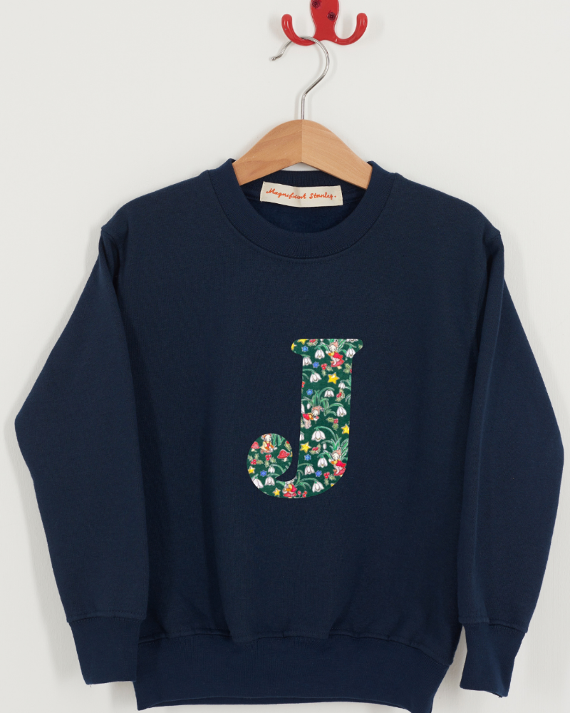 A navy blue crewneck sweatshirt hanging on a wooden hanger against a light gray wall. The sweatshirt features a large floral Liberty Elves patterned letter 'J' on the front.