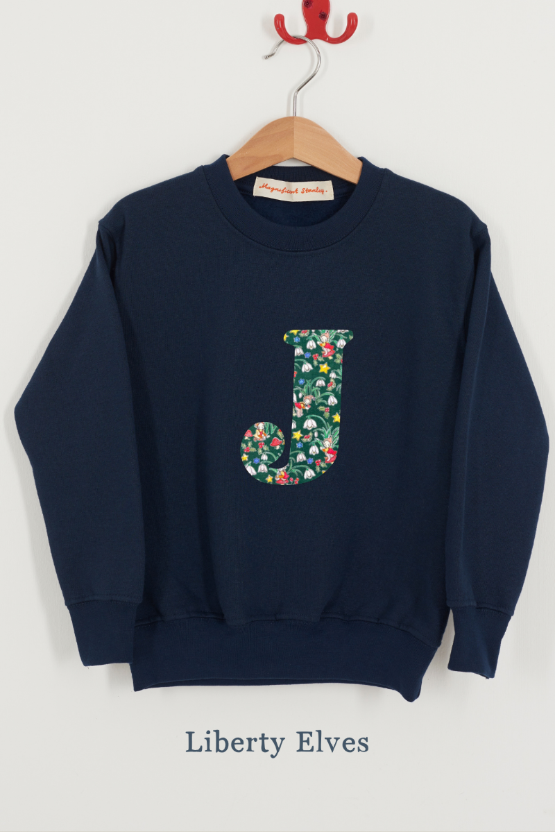 A navy blue crewneck sweatshirt hanging on a wooden hanger against a light gray wall. The sweatshirt features a large floral Liberty Elves patterned letter 'J' on the front.