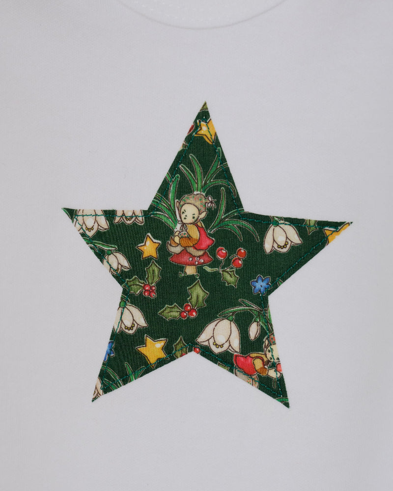 A white long-sleeve baby bodysuit with a green star-shaped patch featuring a festive Liberty Elves print featuring snowdrops, holly, elves and stars, laid flat on a green patterned fabric background. A close up of the appliqué stitching