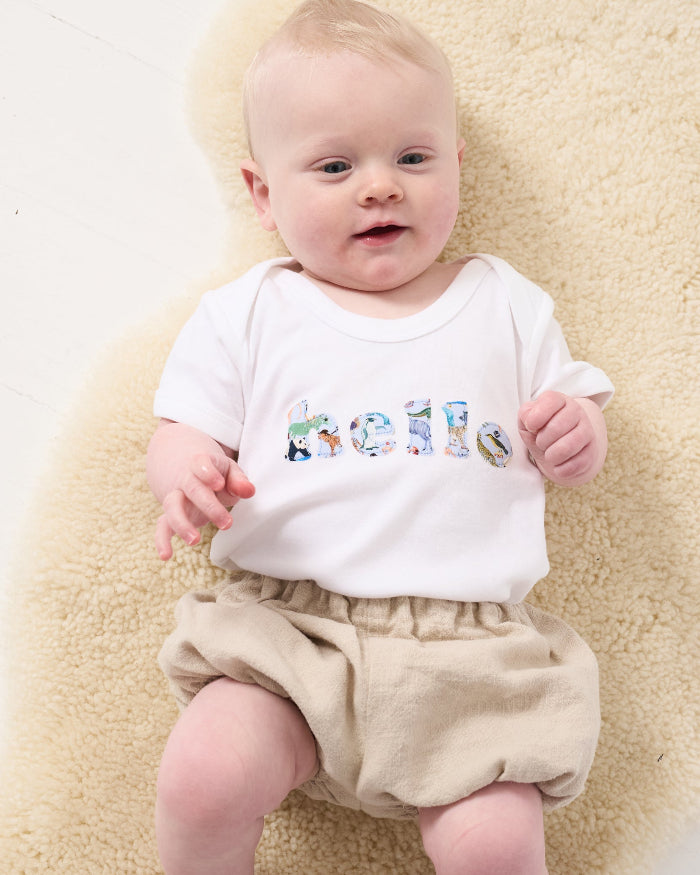 A 3 month old baby wearing a white cotton babygro with the word hello sewn on the front, cut from Quey 2 zoo animal Liberty print.