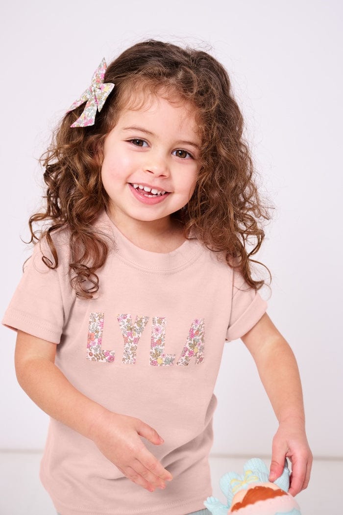 A smiling child wearing a pink t-shirt with her name written in a floral Liberty Print.