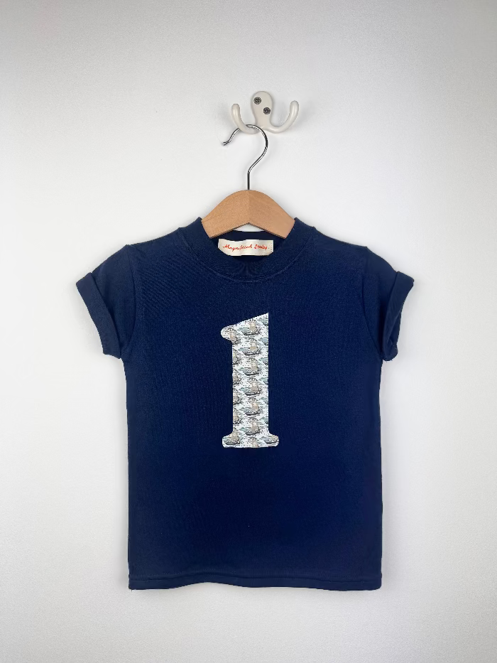 A navy blue short-sleeve kids' t-shirt hanging on a wooden hanger with a white wall hook behind it. The shirt features a large number '1' printed on the chest in a pattern of small gray and white liberty ships.