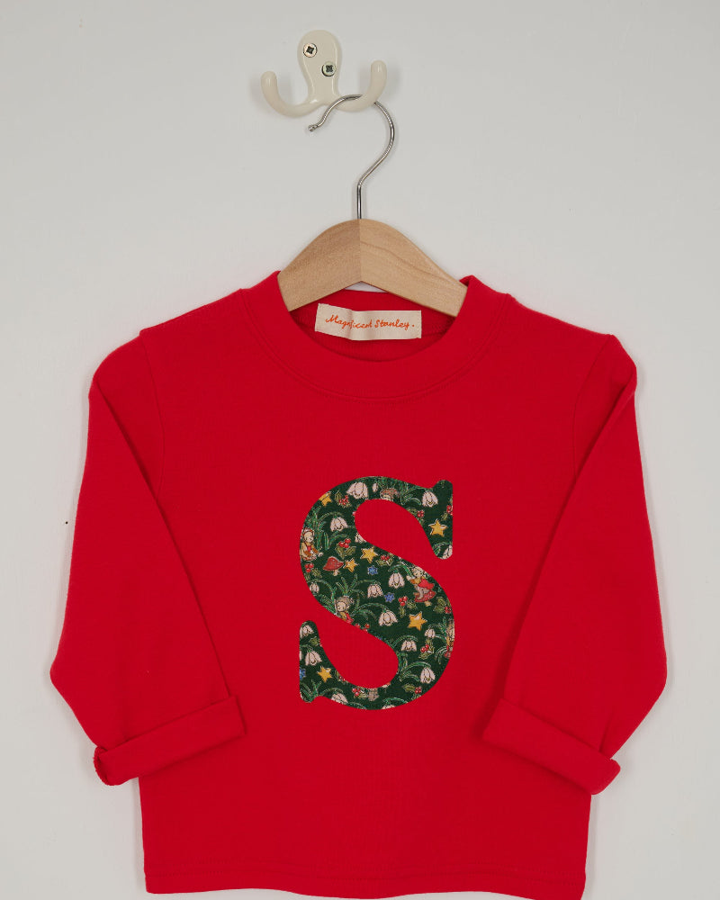 A festive children's outfit set featuring a bright red long-sleeve top with a large green floral monogram letter 'S' on the chest made from Liberty Elves print.