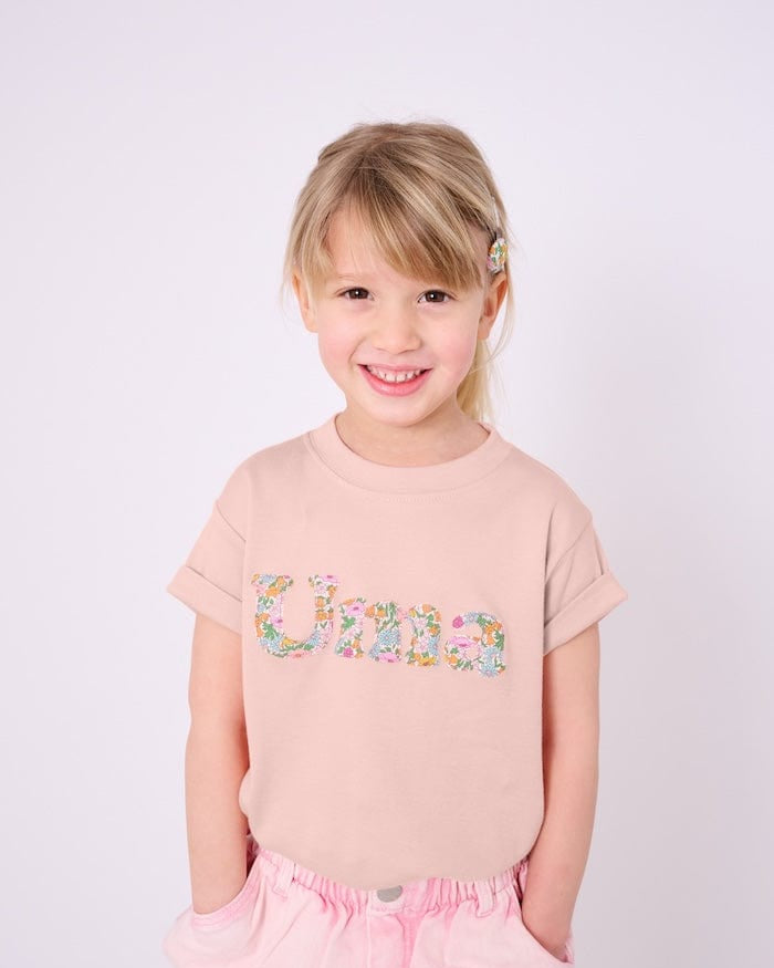 A 4 year old girl wearing a soft pink kids t-shirt with her name cut from Liberty print in lowercase letters.