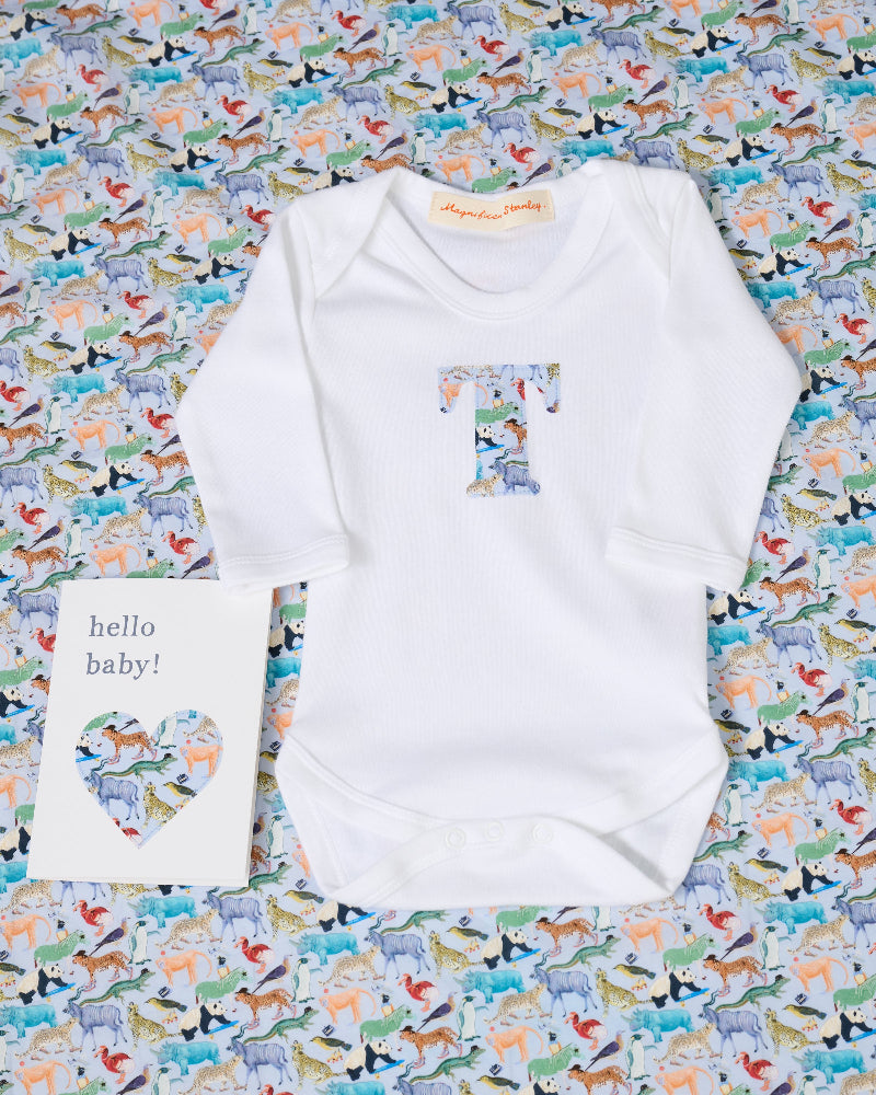 A white long-sleeve baby bodysuit laid flat on a colorful animal-print fabric background, with a large letter T printed on the chest in a pastel multicolor pattern. A small white card reading 'hello baby!' is placed beside it.