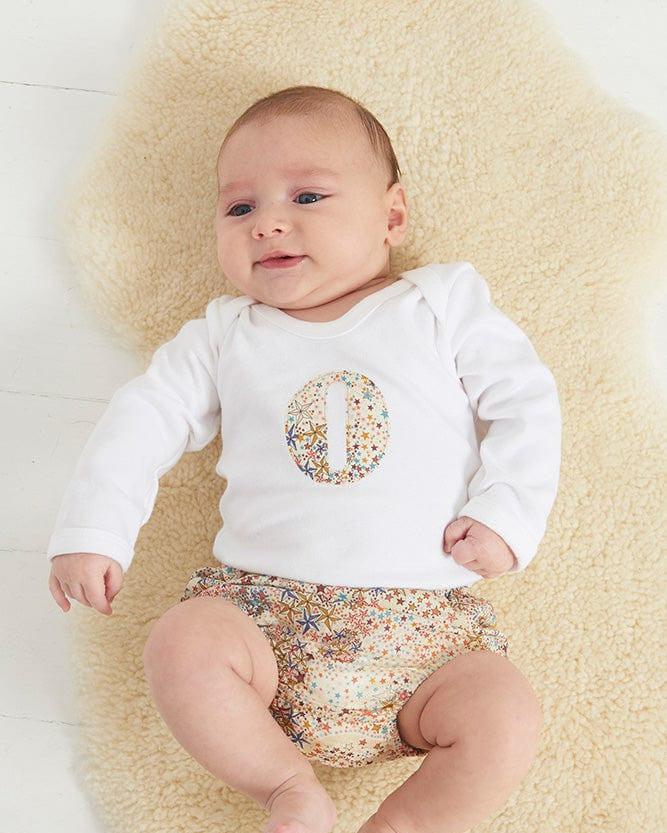 A smiling newborn baby wearing a personalised white bodysuit with an initial on the front cut from Liberty Adelajda starry fabric.
