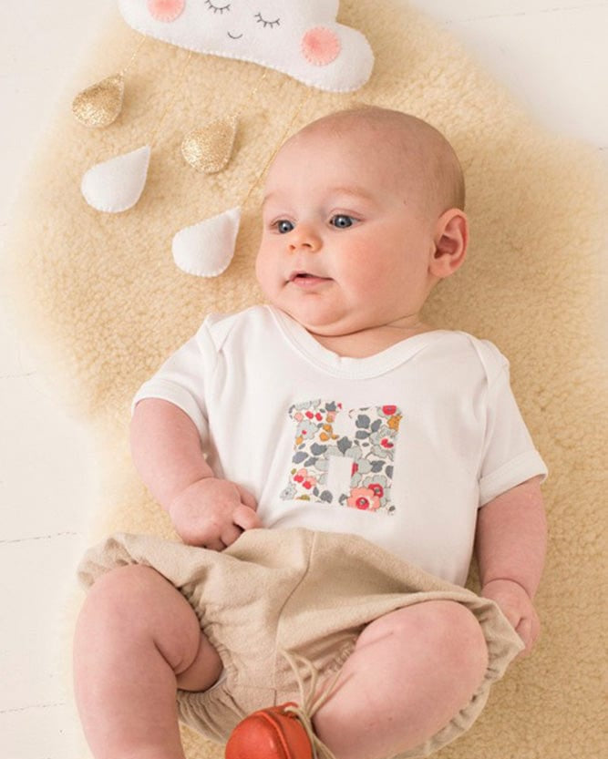 A smiling newborn baby wearing a personalised white bodysuit with an initial on the front cut from betsy grey Liberty floral fabric.