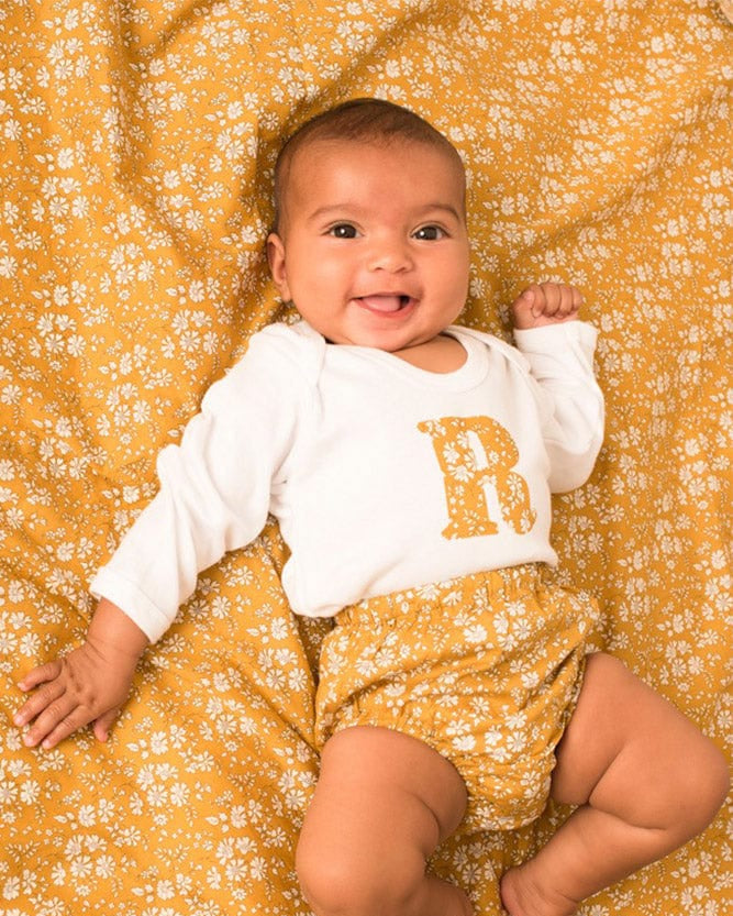 A smiling newborn baby wearing a personalised white bodysuit with an initial on the front cut from Capel Liberty floral fabric.