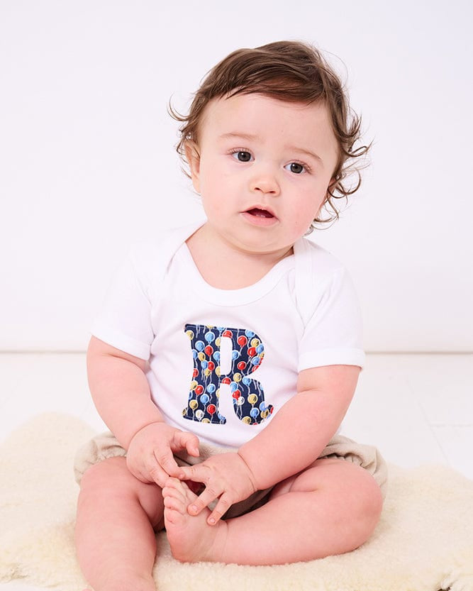 A smiling newborn baby wearing a personalised white bodysuit with an initial on the front cut from Ethan's party balloon Liberty balloon fabric.