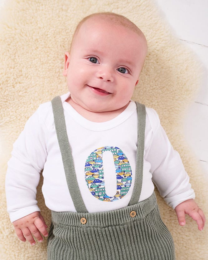 A smiling newborn baby wearing a personalised white bodysuit with an initial on the front cut from Liberty cars fabric.