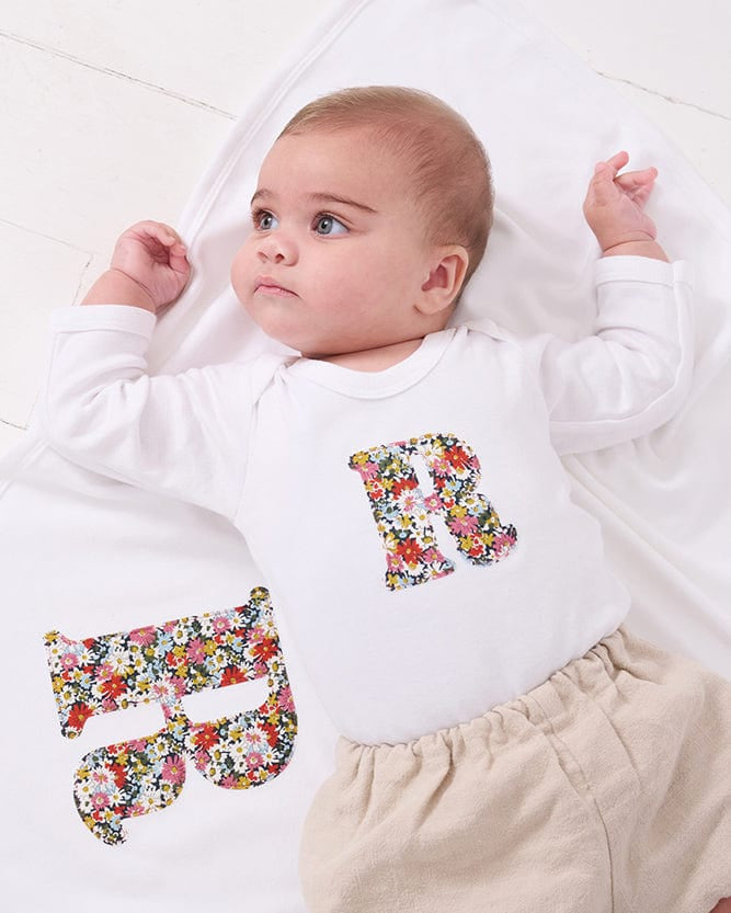A smiling newborn baby wearing a personalised white bodysuit with an initial on the front cut from Liberty floral fabric. She is laying on a matching personalised blanket.
