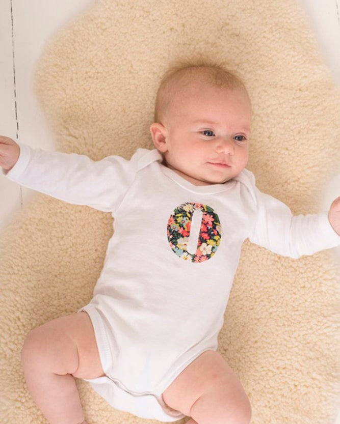 A smiling newborn baby wearing a personalised white bodysuit with an initial on the front cut from Liberty floral fabric.