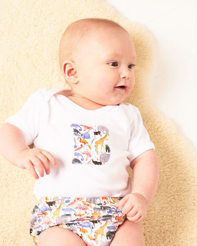 A smiling newborn baby wearing a personalised white bodysuit with an initial on the front cut from Liberty zoo animal fabric.