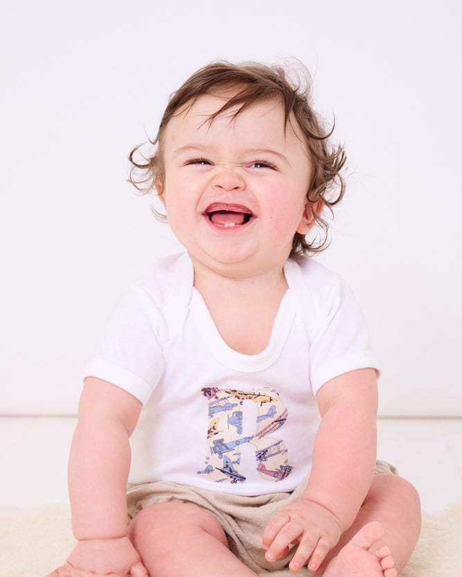 A smiling newborn baby wearing a personalised white bodysuit with an initial on the front cut from Liberty jet plane fabric.
