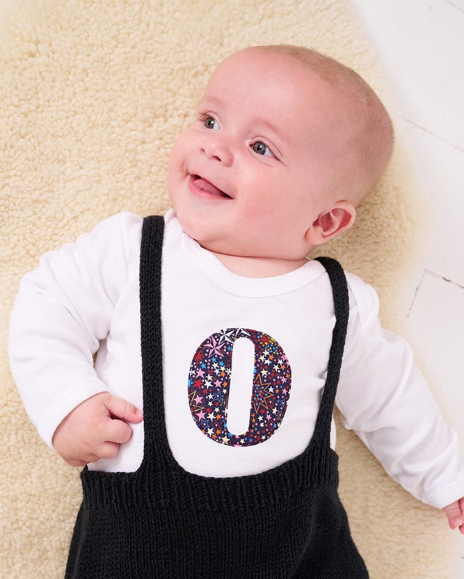A smiling newborn baby wearing a personalised white bodysuit with an initial on the front cut from Liberty Adelajda starry fabric.