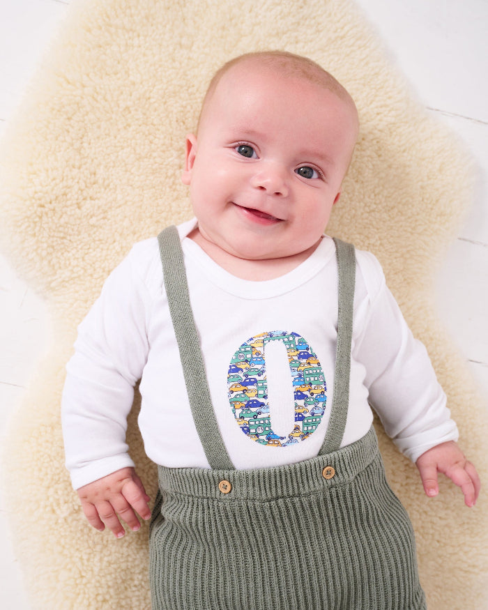 A baby wearing a Personalised Bodysuit in hop on hop off Liberty Print featuring vintage cars and busses.