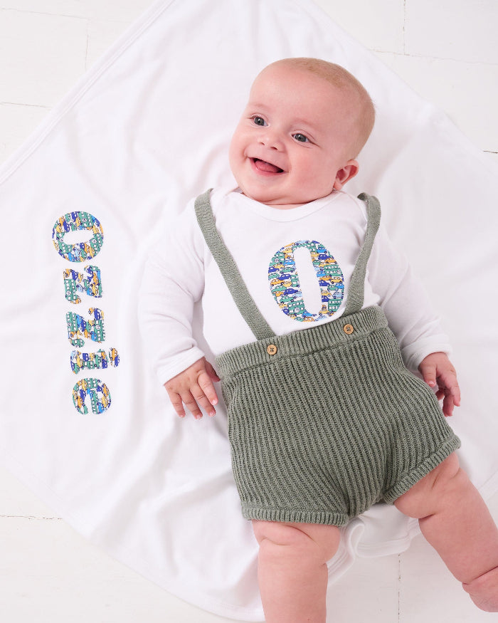 A baby wearing a Personalised Bodysuit in Hop on hop off Liberty Print featuring cars. The baby is laying on a personalised cotton blanket with his name on
