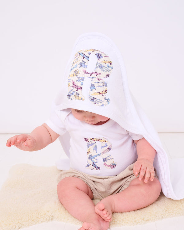 A 6 month old baby boy wearing a Personalised Bodysuit in Tom's Jet Liberty Print with his initial sewn onto the front. He is also wearing a personalised hooded towel.