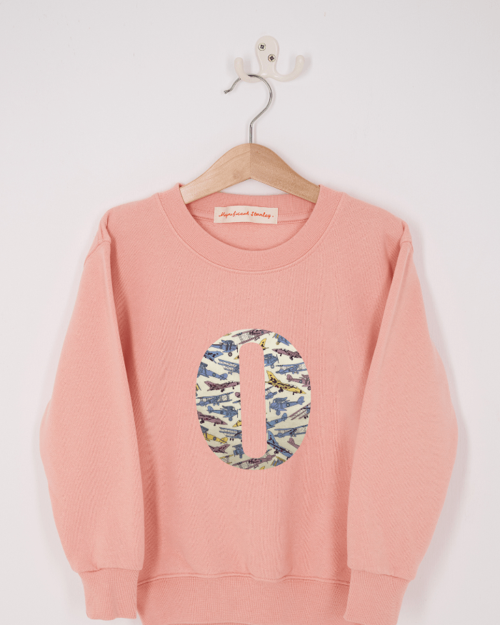 A pink personalised sweatshirt with an initial sewn on to the front. The initial is made from liberty print featuring vintage jet planes.