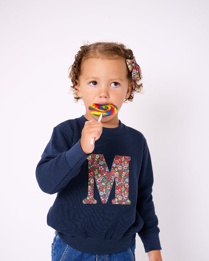 A 3 year old girl wearing a  navy Magnificent Stanley personalised Navy Sweatshirt with her initial on the front in floral emma louise liberty print.