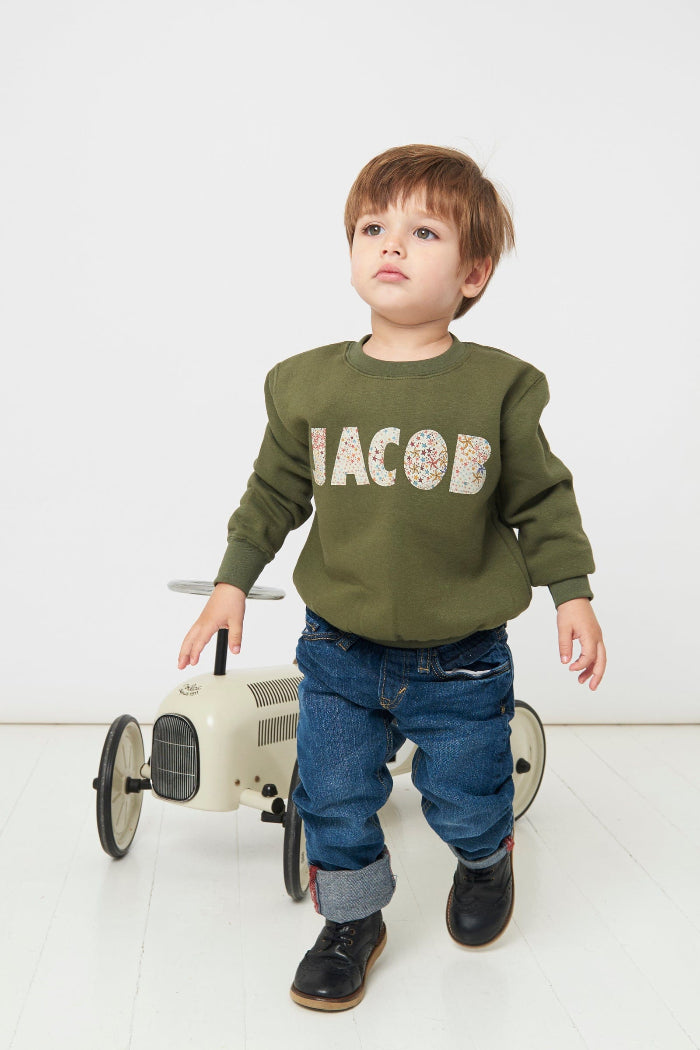 A 4 year old boy wearing a Magnificent Stanley khaki sweatshirt with his name 'Jacob' sewn on the front in a starry adelajda Liberty Print