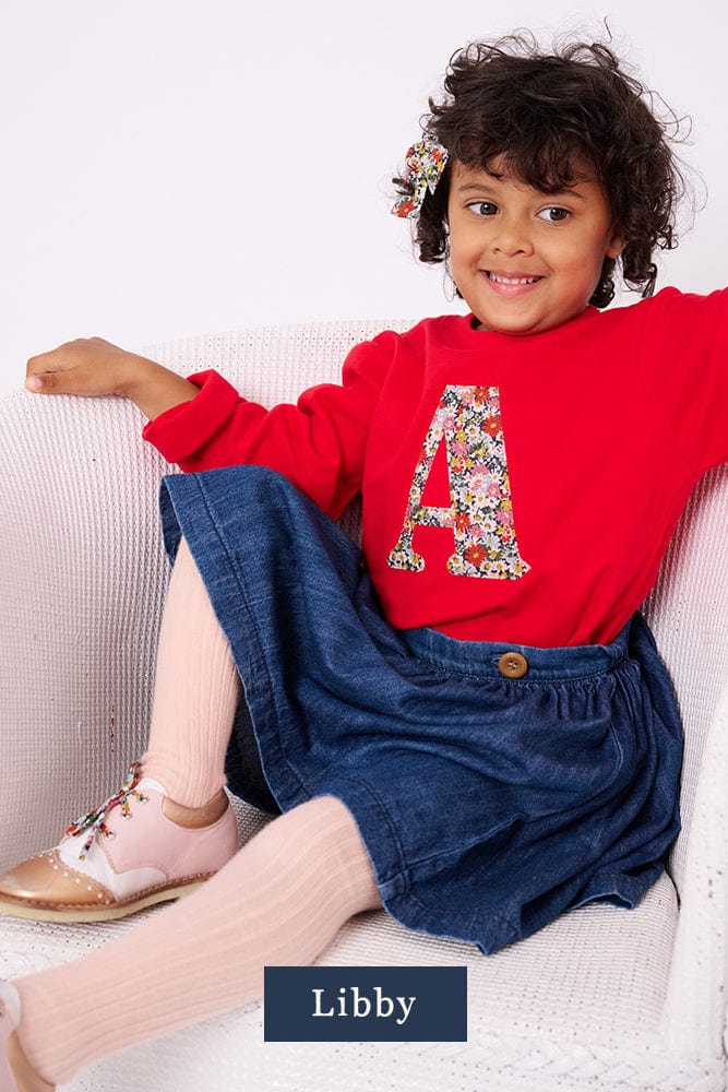 A 3 year old girl wearing a red personalised t-shirt with a liberty floral letter A appliquéd on the front.