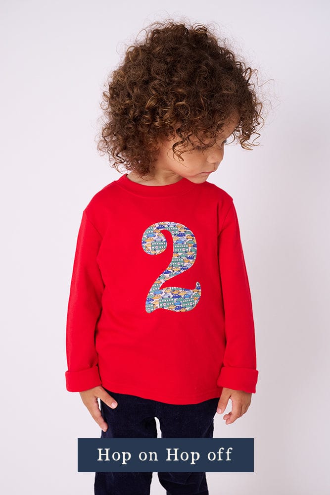 A 2 year old boy wearing a red 2nd birthday t-shirt with a liberty cars print number 2 appliquéd on the front.