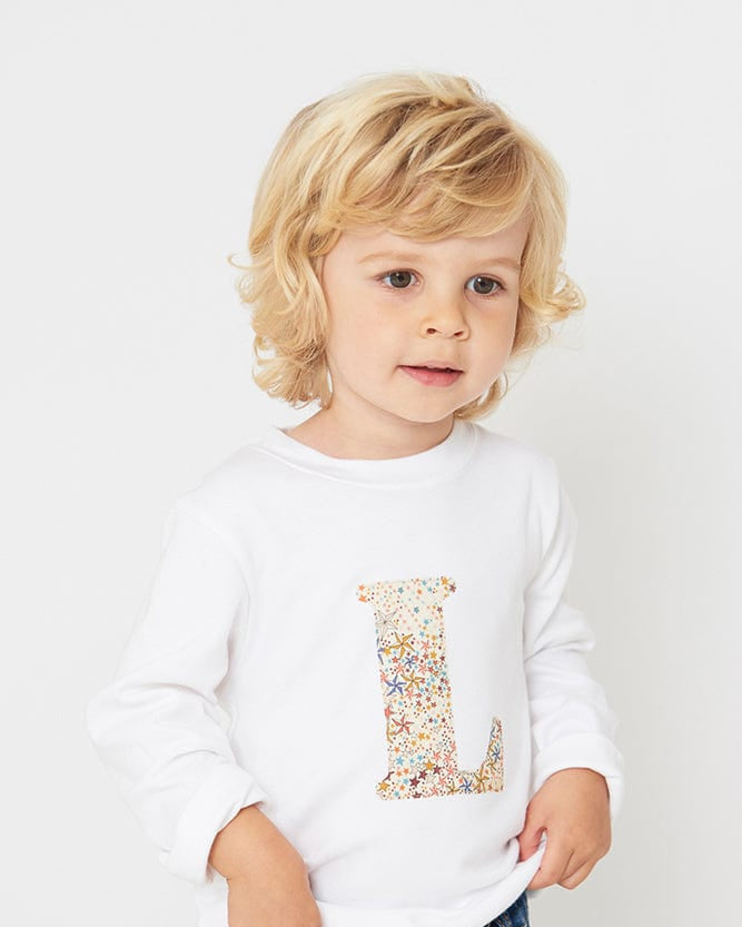 A 3 year old boy wearing a personalised white cotton t-shirt with her initial on in Adelajda star Liberty fabric.
