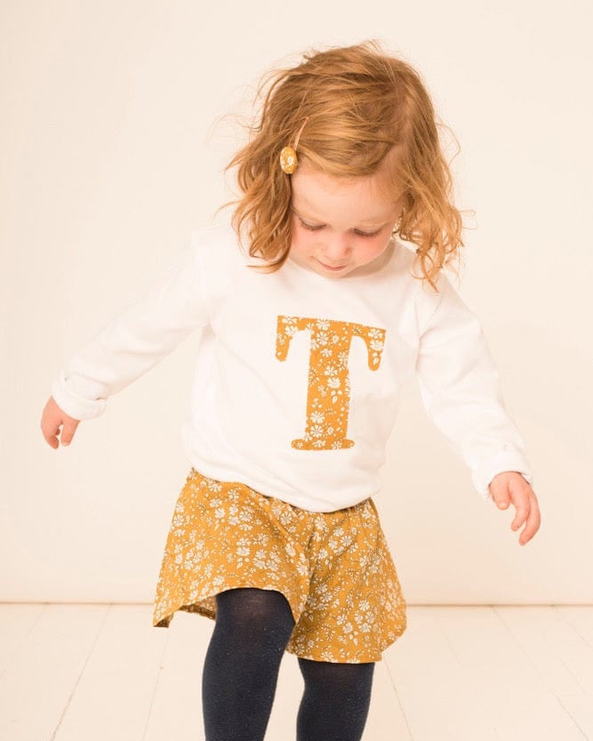 A 2 year old girl wearing a personalised white cotton t-shirt with her initial on in Capel floral Liberty fabric.