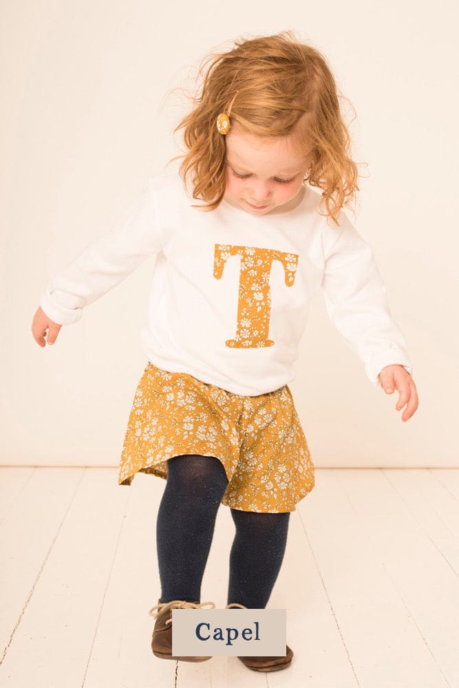 A 2 year old girl wearing a personalised white cotton t-shirt with her initial on in Capel floral Liberty fabric.