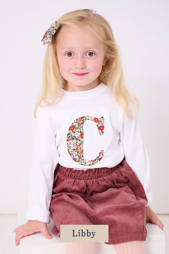 A 4 year old girl wearing a personalised white cotton t-shirt with her initial on in Libby floral Liberty fabric.