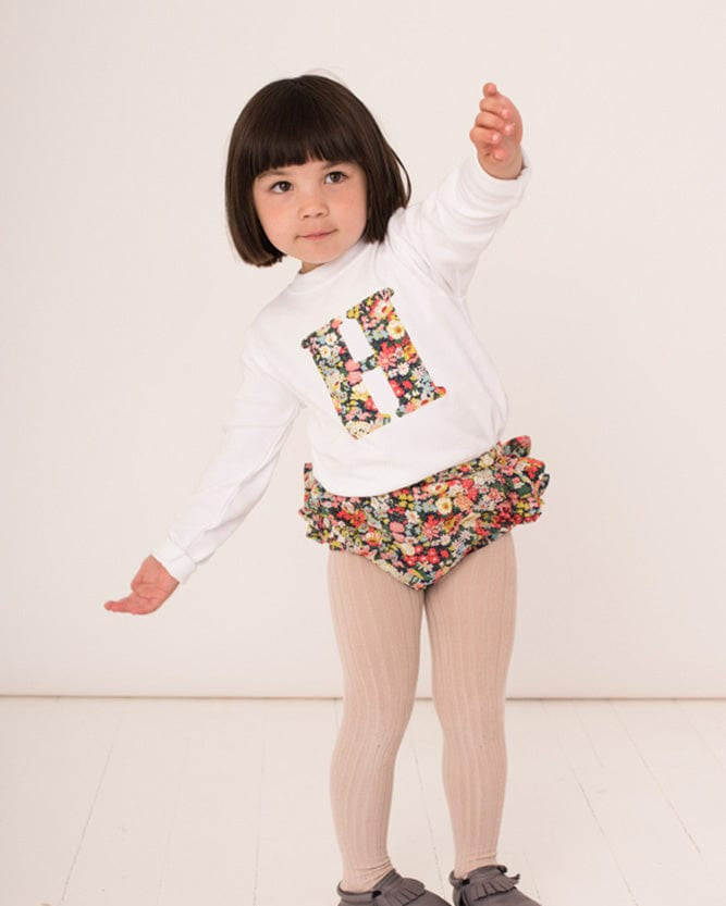 A 2 year old girl wearing a personalised white cotton t-shirt with his initial on in Thorpe floral Liberty fabric.