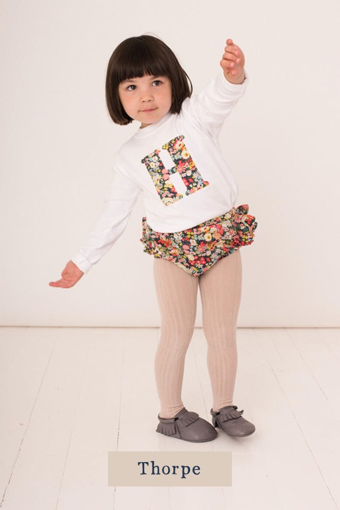 A 2 year old girl wearing a personalised white cotton t-shirt with his initial on in Thorpe floral Liberty fabric.