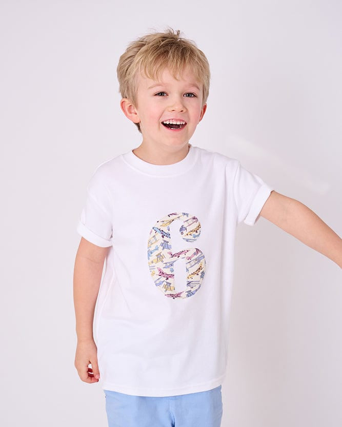 A 6 year old boy wearing a birthday white cotton t-shirt with the number 6 on in jet planes Liberty fabric.