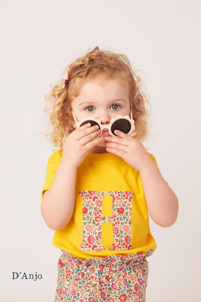 a smiling 2 year old girl wearing a personalised kids yellow t-shirt with an initial cut from d'anjo liberty floral print on the front