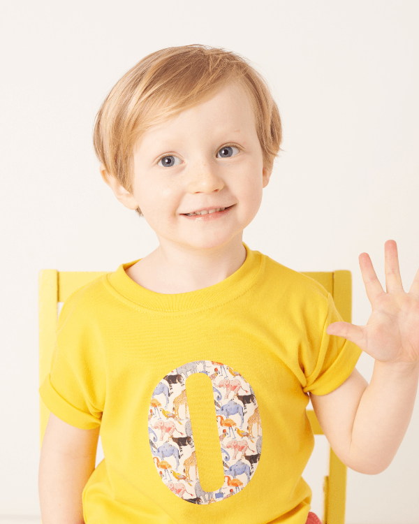 a smiling 3 year old boy wearing a personalised kids yellow t-shirt with an initial cut from queue for the zoo liberty animal print on the front