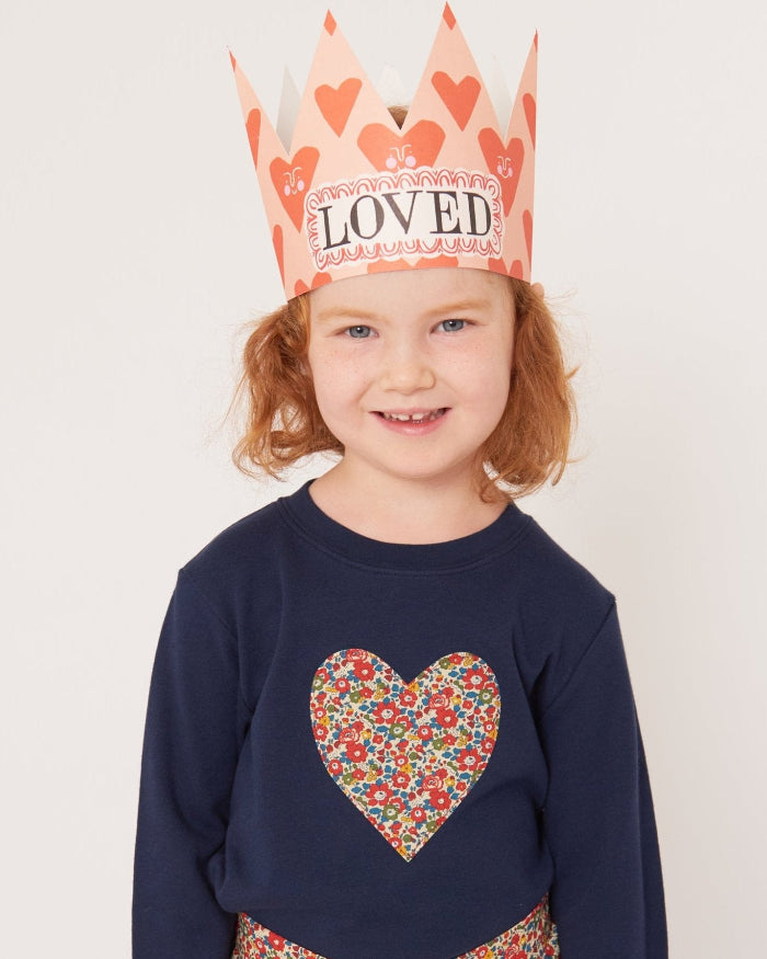 A 4 year old girl wearing a children's valentines outfit. A navy cotton t-shirt with a Liberty floral heart sewn on the front and a crown that says 'loved' on it.