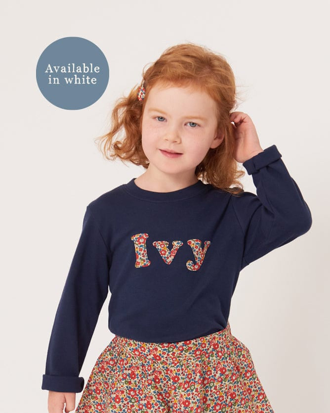 A 4 year old girl wearing a personalised navy cotton t-shirt with her name on the front. Her name is appliquéd in lowercase letters in Liberty floral fabric.