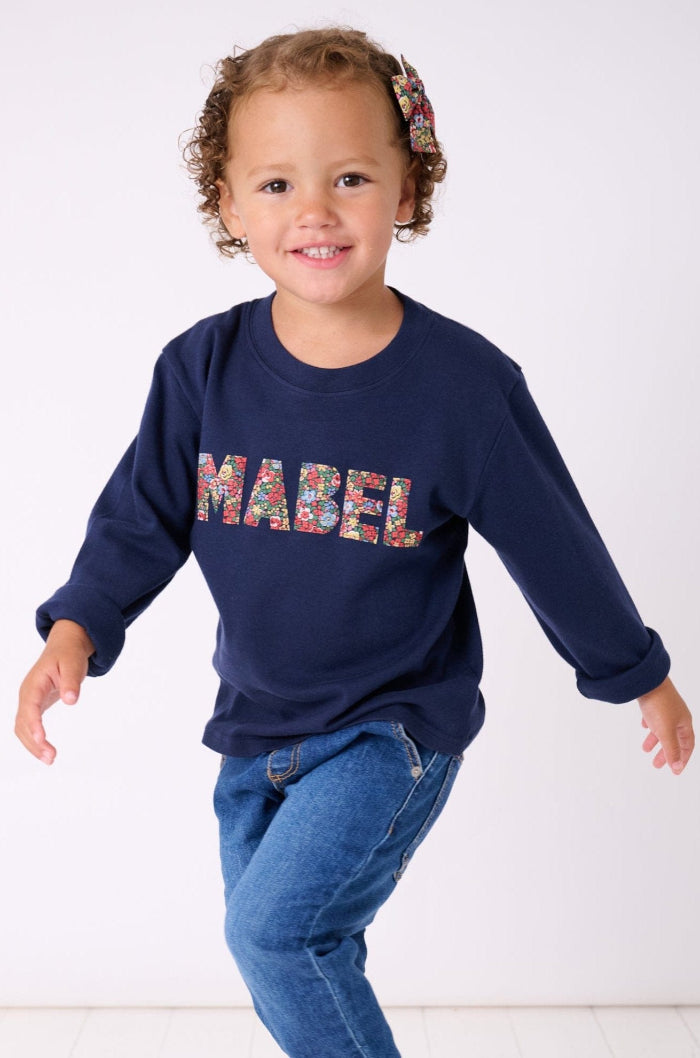 A smiling 3 year old girl wearing a navy cotton personalised t-shirt with her name on cut from floral liberty print.