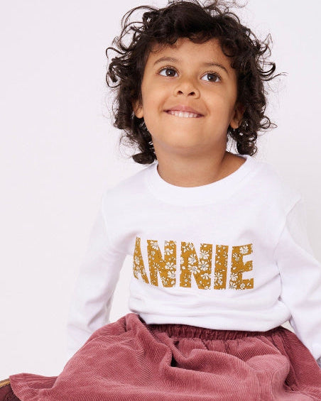 A young girl wearing a white personalised t-shirt with her name on the front cut from Capel Mustard liberty print.