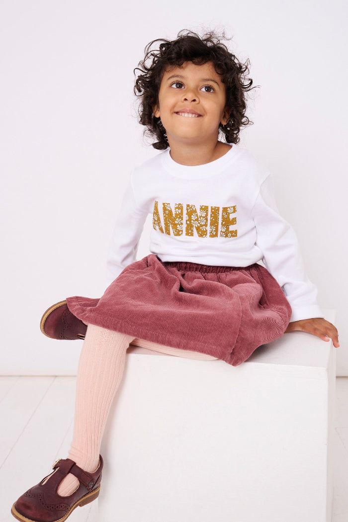 A young girl wearing a white personalised t-shirt with her name on the front cut from Capel Mustard liberty print.