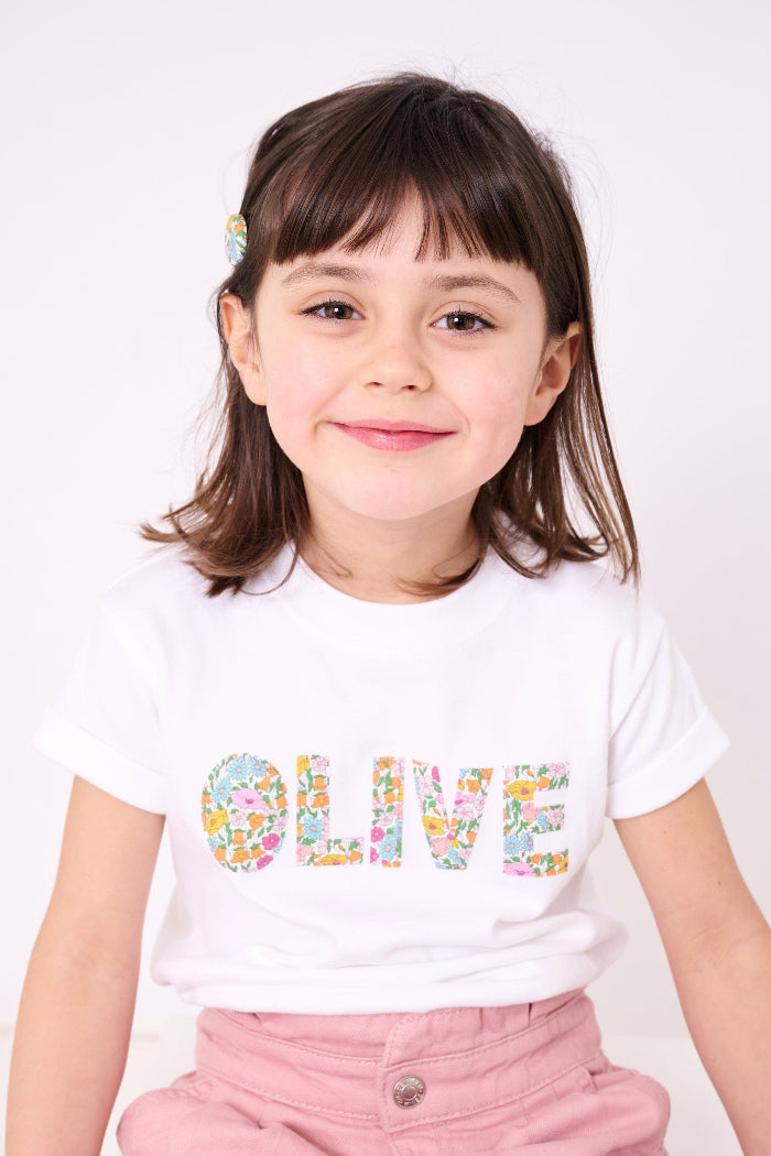 A young girl wearing a white personalised t-shirt with her name on the front cut from Poppy Forest liberty print.