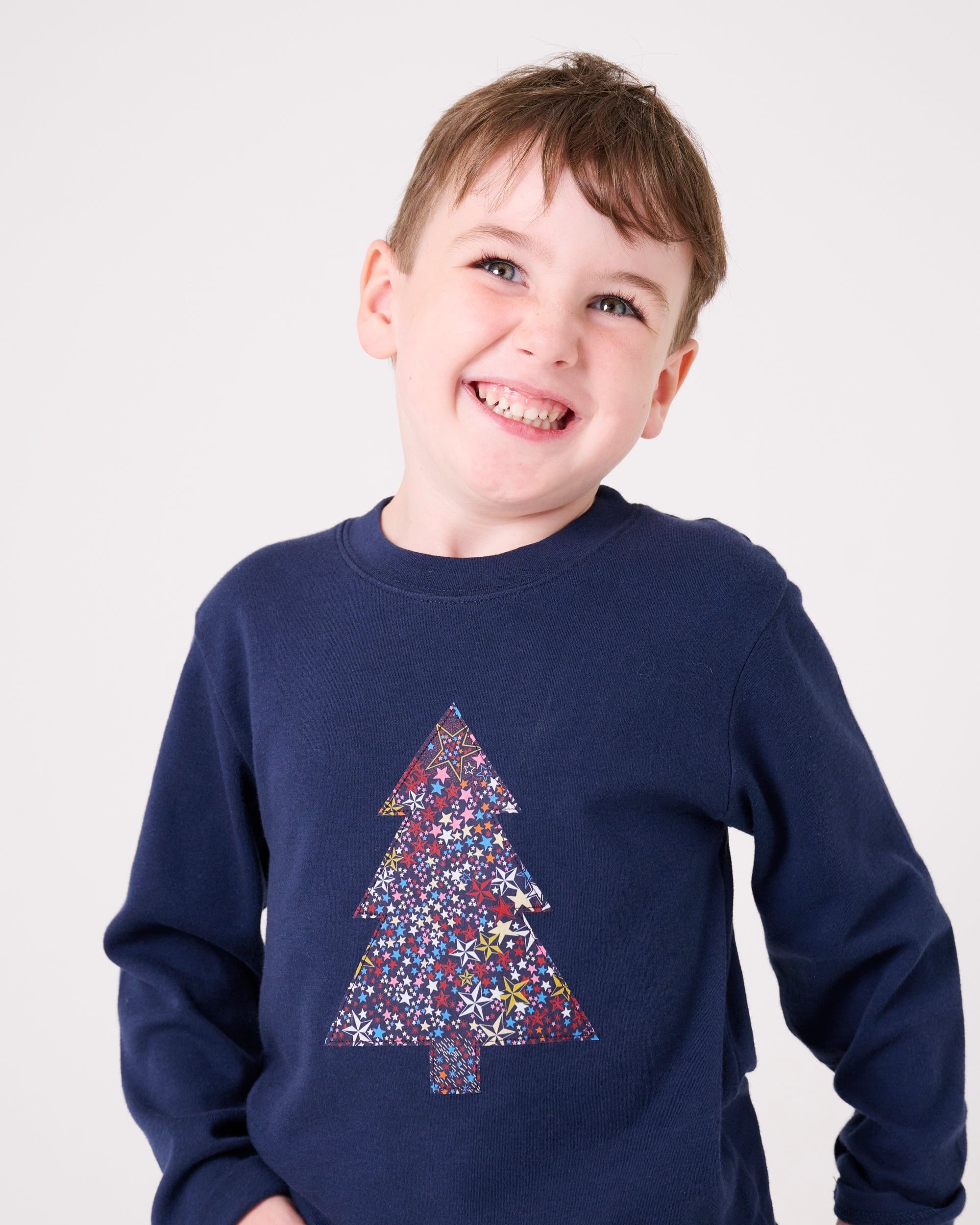 A 3 year old boy wearing a navy long sleeve Christmas t-shirt with a Christmas tree on the front made from Liberty Adelajda's Wish star print on the front.