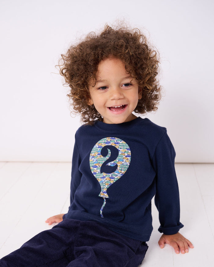 A 2 year old birthday boy wearing a navy t-shirt with a number 2 in a balloon on the front. The balloon is cut from Hop on hop off cars Liberty print.