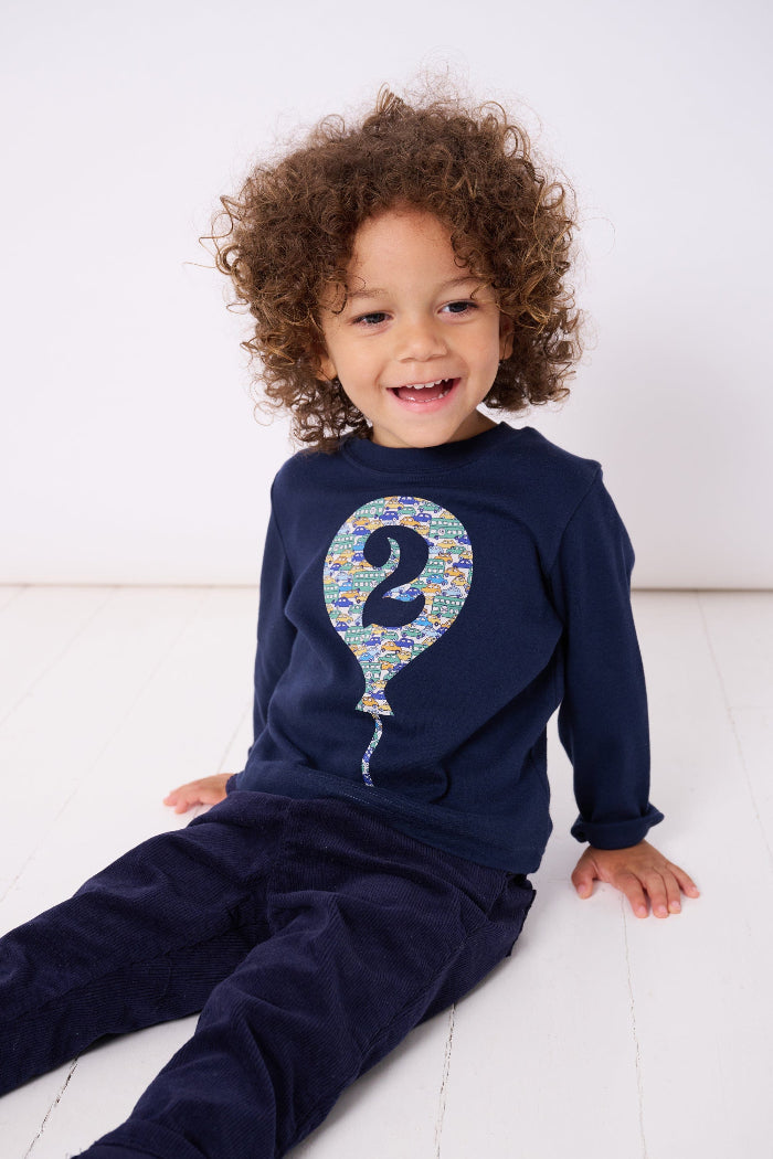 A 2 year old birthday boy wearing a navy t-shirt with a number 2 in a balloon on the front. The balloon is cut from Hop on hop off cars Liberty print.