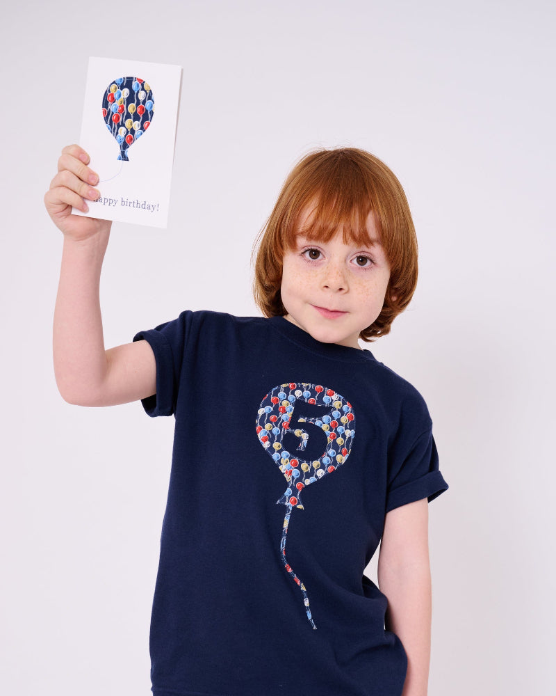 A 5 year old birthday boy wearing a navy t-shirt with a number 5 in a balloon on the front. The balloon is cut from Ethan's Party balloon Liberty print.