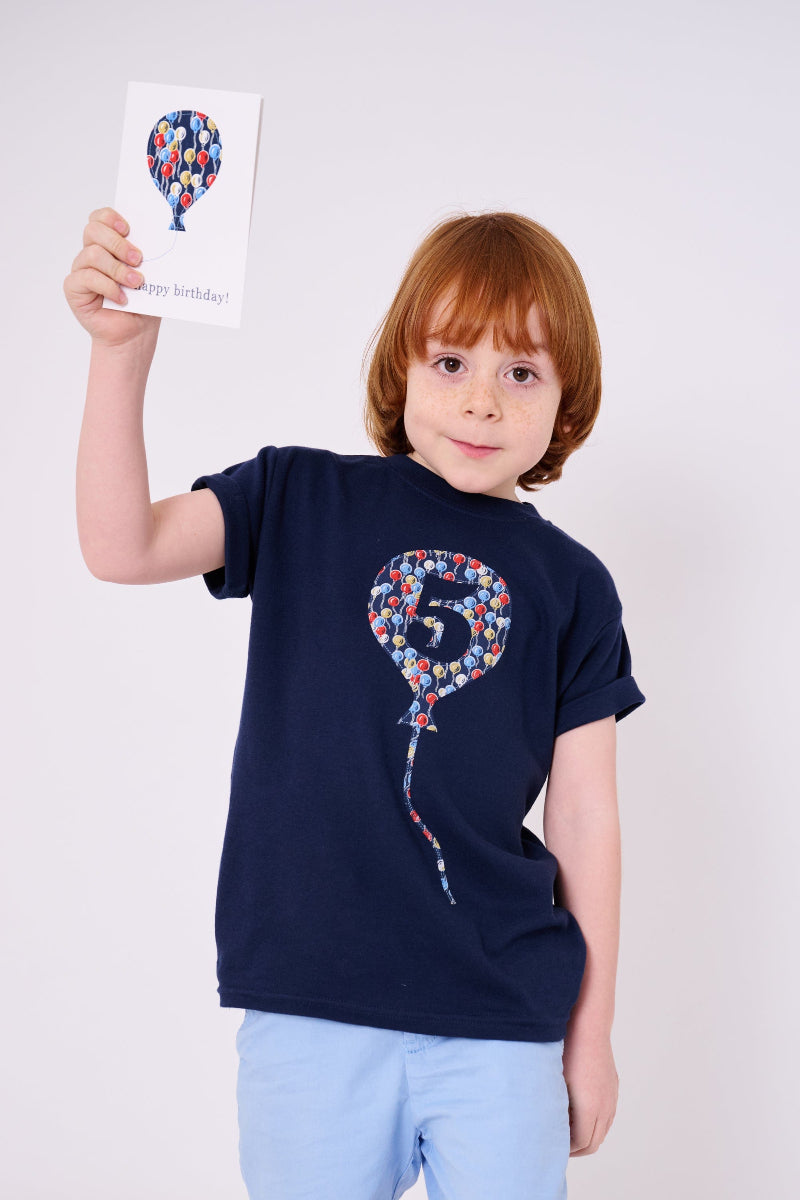 A 5 year old birthday boy wearing a navy t-shirt with a number 5 in a balloon on the front. The balloon is cut from Ethan's Party balloon Liberty print.