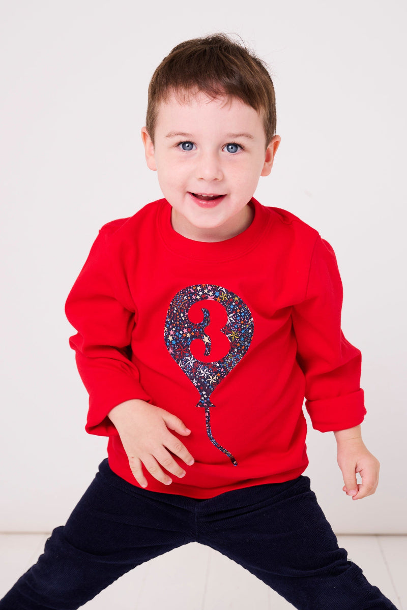 A 3 year old wearing a red cotton t-shirt with a liberty star print birthday balloon on the front with a number 3 on it.